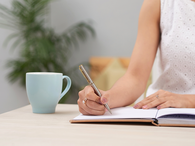 mujer-irreconocible-en-vestido-sentado-en-el-interior-en-el-escritorio-y-escribiendo-en-el-diario