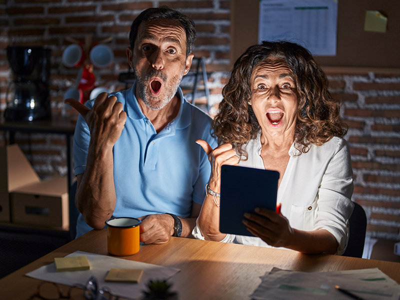 una-pareja-hispana-de-mediana-edad-usando-el-touchpad-sentada-en-la-mesa-por-la-noche-sorprendida-senalando-con-el-dedo-de-la-mano-hacia-el-lado-boca-abierta-expresion