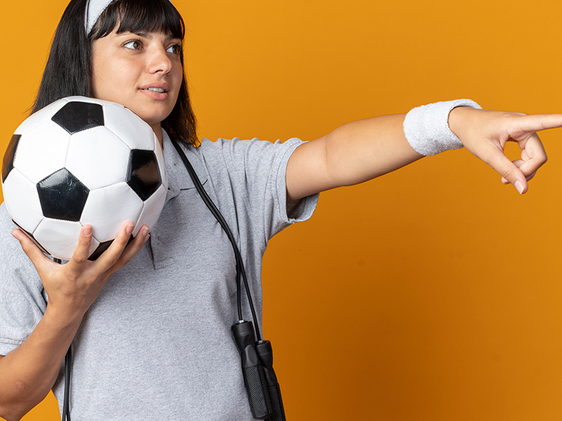 chica-joven-fitness-con-diadema-con-cuerda-alrededor-del-cuello-sosteniendo-un-balon-de-futbol-mirando-un-lado-con-una-sonrisa-en-la-cara-apuntando-con-el-dedo-in